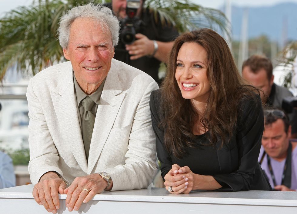 clint eastwood and angelina jolie lean against a white ledge and stands outside while smiling for photos, he wears a light colored suit jacket and gray collared shirt and tie, she wears a black outfit and jacket