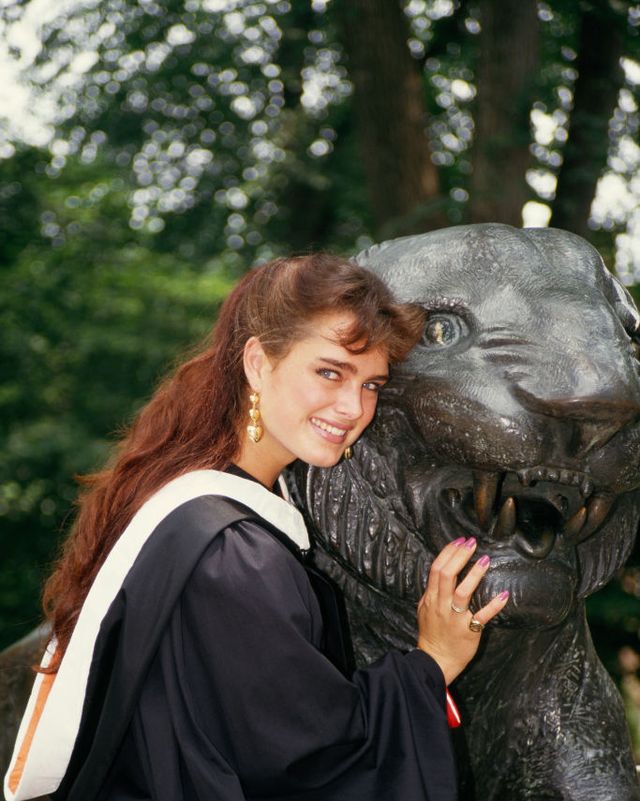 brooke shields smiles at the camera while posing with a metal tiger statue, she wears a black graduation gown with a white neck sash, large gold earrings, and two rings on her hand