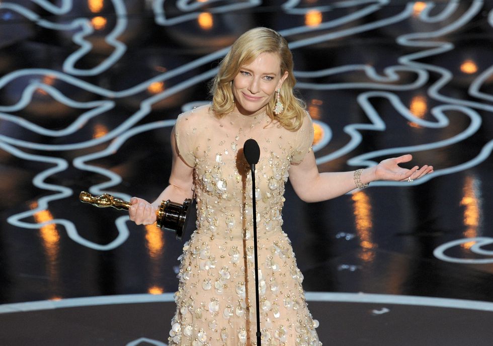 cate blanchett wearing a beige dress, standing in front of a microphone on a stage, holding an oscar statuette and gesturing with her left arm