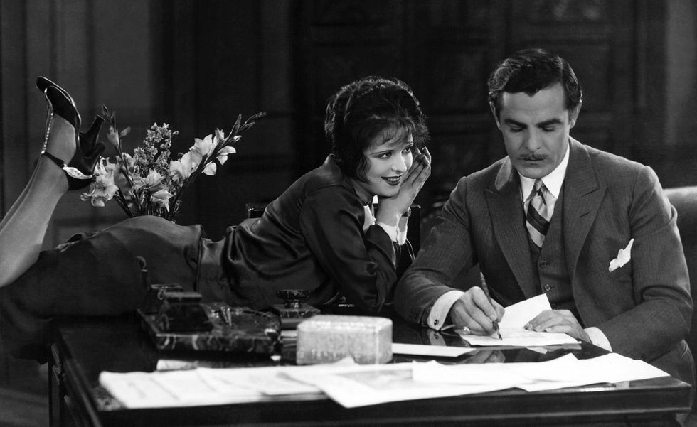 clara bow and antonio moreno in a scene from the movie it, she lies on her stomach with her legs up in the air and smiles at him, he sits at a table writing, several other papers are on the table as well