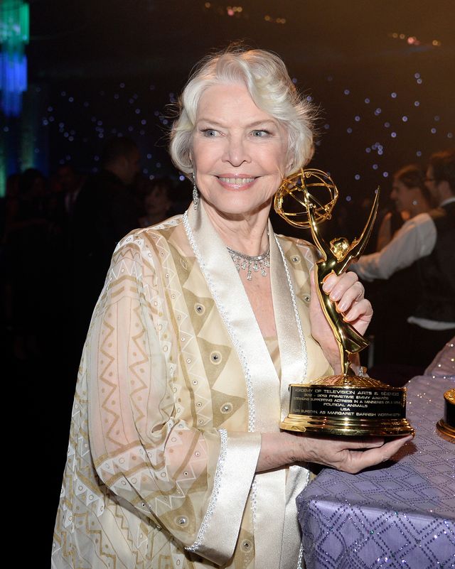 ellen burstyn smiles and looks to the left of the camera, she holds her emmy award while standing next to a table with a purple tablecloth, she wears a cream, tan and gold caftan