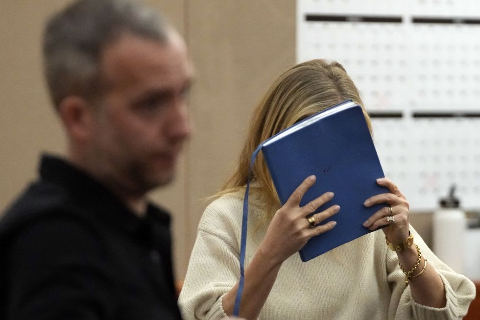 gwyneth paltrow, wearing a white sweater, holds a blue book in front of her face in a courtroom, as a man in a black shirt stands in the foreground