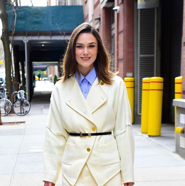 kiera knightley smiles at the camera, she stands on a city sidewalk wearing a white suit with a black waist belt and blue collared shirt