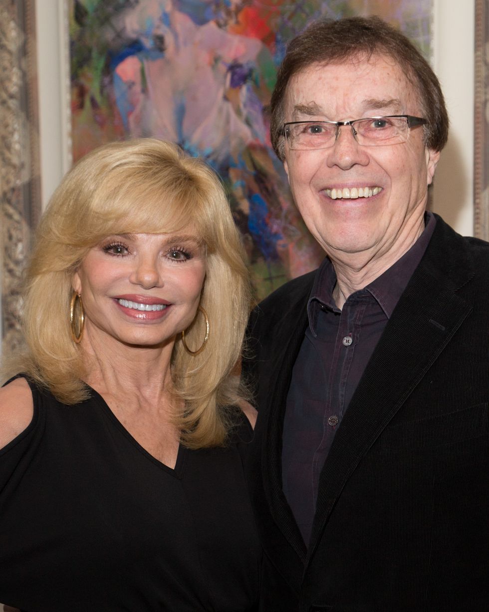 a woman and a man stand together and smile for a photo, she wears a black dress with gold earrings, he wears a black suit coat and a dark collared shirt with glasses