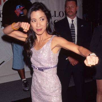 michelle yeoh, wearing a pink dress and pink high heel shoes, makes a fighting pose while standing on a red carpet at a movie premiere
