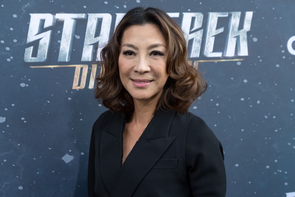 michelle yeoh smiling, wearing a black shirt, standing in front of a backdrop with stars and the words star trek discovery on them