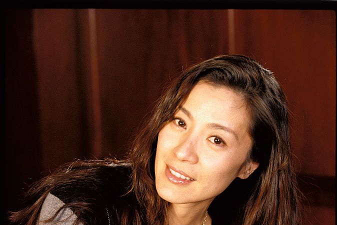 michelle yeoh smiling into the camera and sitting cross legged, wearing a black shirt