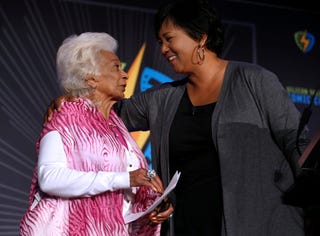 nichelle nichols and mae c jemison look at one another while standing together and speaking, nichols wears a pink vest over a white long sleeve sweater, jemison has one arm around nichols shoulders and wears a gray sweater and a black top