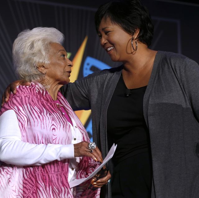 nichelle nichols and mae c jemison look at one another while standing together and speaking, nichols wears a pink vest over a white long sleeve sweater, jemison has one arm around nichols shoulders and wears a gray sweater and a black top