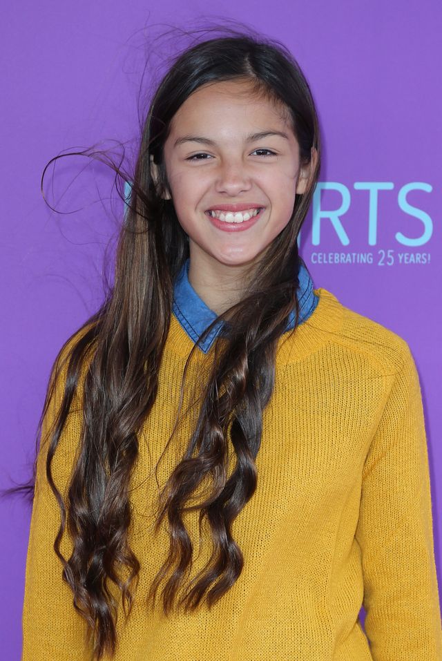 olivia rodrigo smiles for a photo in a golden yellow sweater over a blue collared shirt