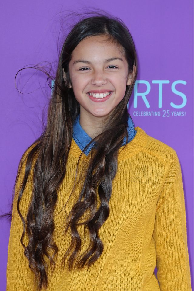 olivia rodrigo smiles for a photo in a golden yellow sweater over a blue collared shirt
