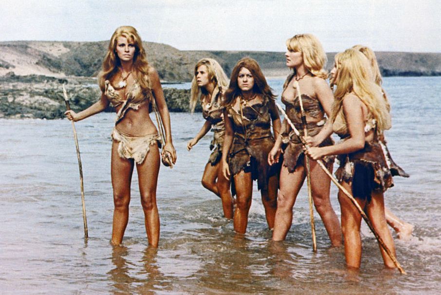 five cavewomen wearing tattered fur skin clothing stand in ankle high water, with several of them clutching spears