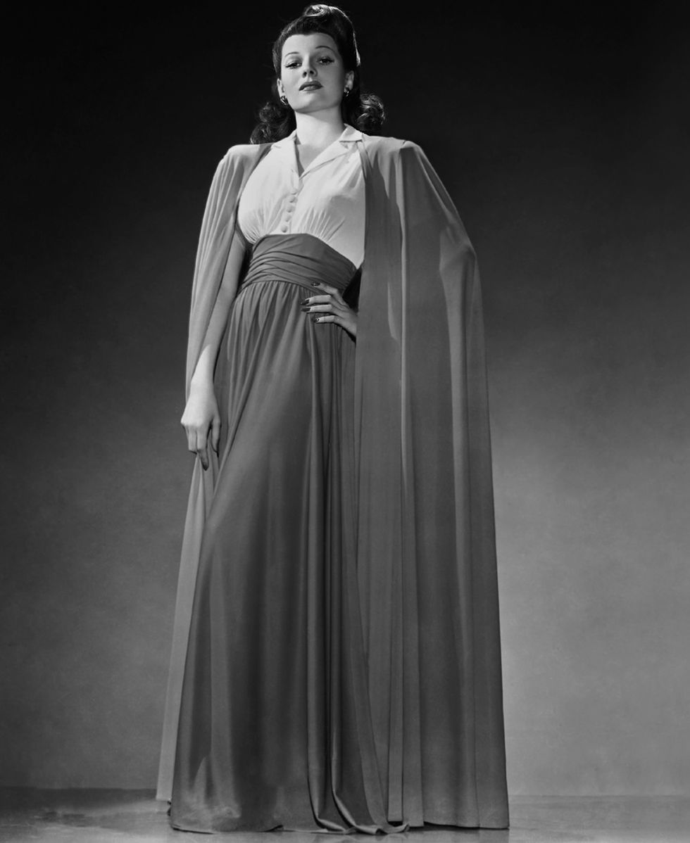 rita hayworth stands and looks directly at the camera, she wears a floor length skirt, light colored blouse and floor length cape