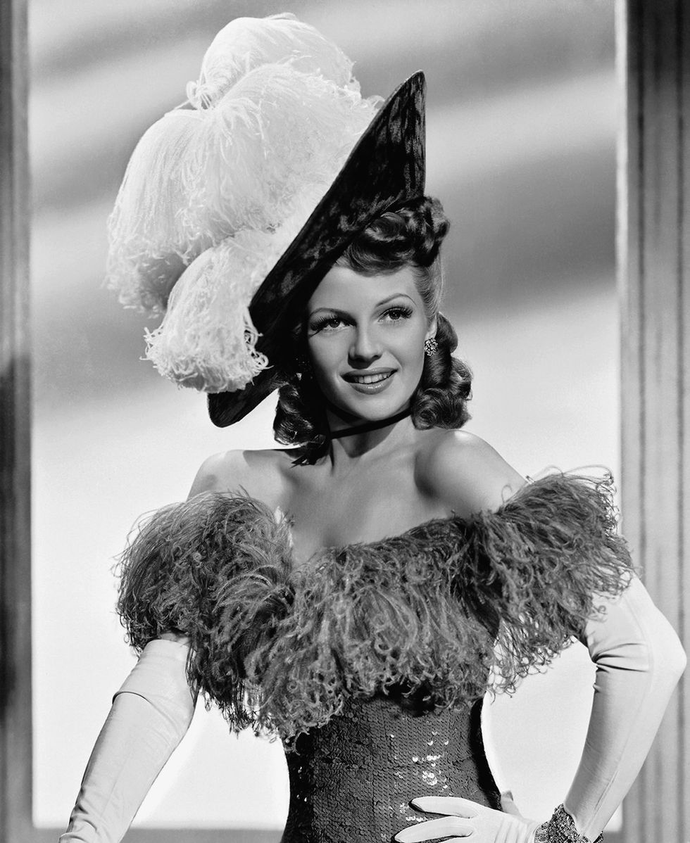 rita hayworth poses with one hand on her hip, she wears a sequin gown with a large feather trim, a matching hat and white gloves