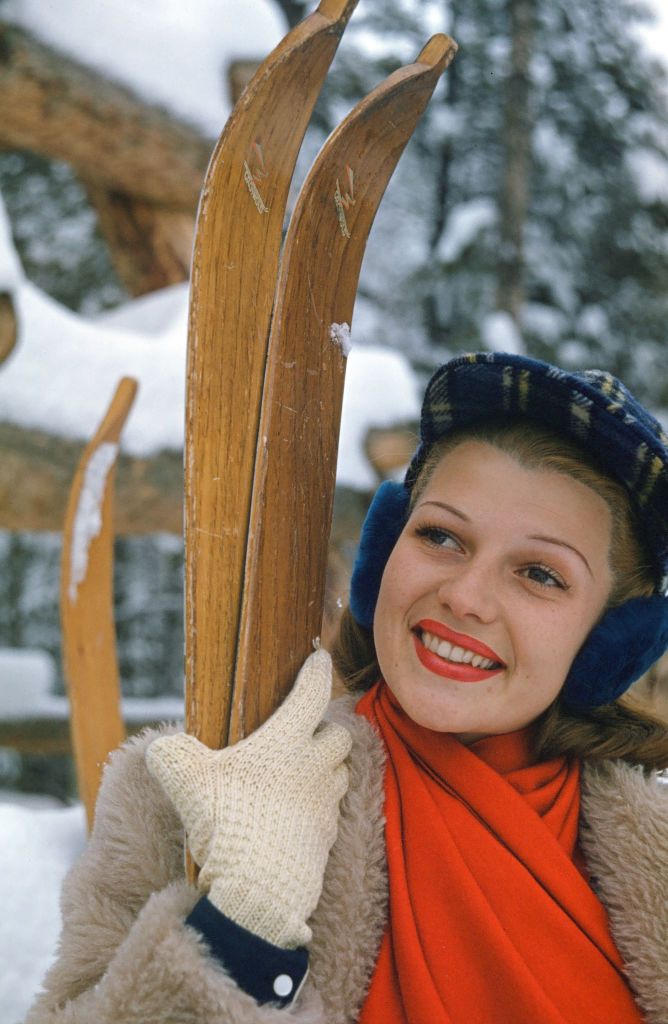 rita hayworth smiles and looks left of the camera, she wears a tan fleece jacket, orange scarf, and blue plaid hat and holds two wooden skis