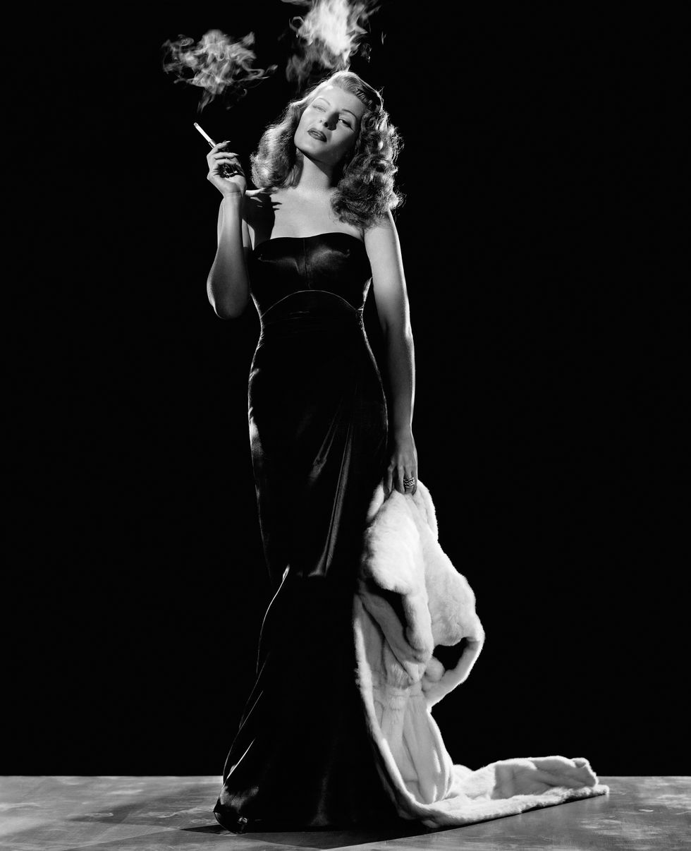 rita hayworth smokes a cigarette and stands in a dark strapless dress, she holds a fur jacket by her leg