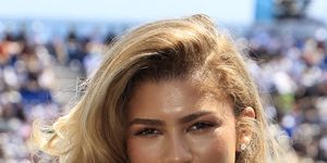zendaya smiles at the camera, she wears a white sweater and stud earrings, blurry crowds of people fill stands behind her