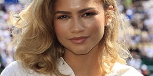 zendaya smiles at the camera, she wears a white sweater and stud earrings, blurry crowds of people fill stands behind her