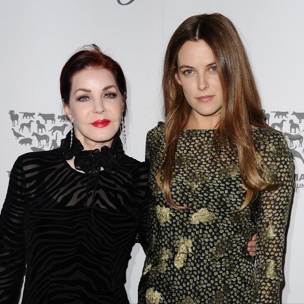 priscilla presley and riley keough pose for a photo together and smile at the camera, presley wears a black sheer dress, keough wears a green and black patterned long sleeve dress
