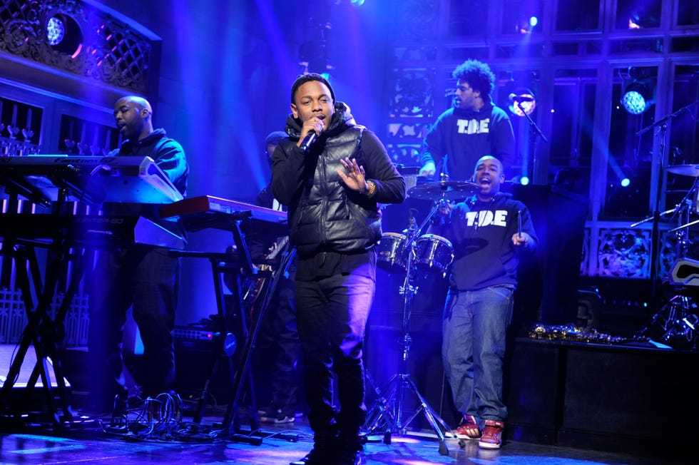 kendrick lamar stands on a stage flooded in blue light and sings into a wireless microphone he holds, three men play instruments in the background