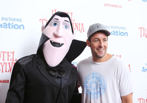adam sandler, wearing a white t shirt and gray hat, stands smiling next to a costumed version of his vampire character from the film hotel transylvania