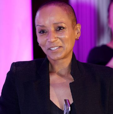 a woman standing and slightly smiling, wearing a black suit with close cropped hair