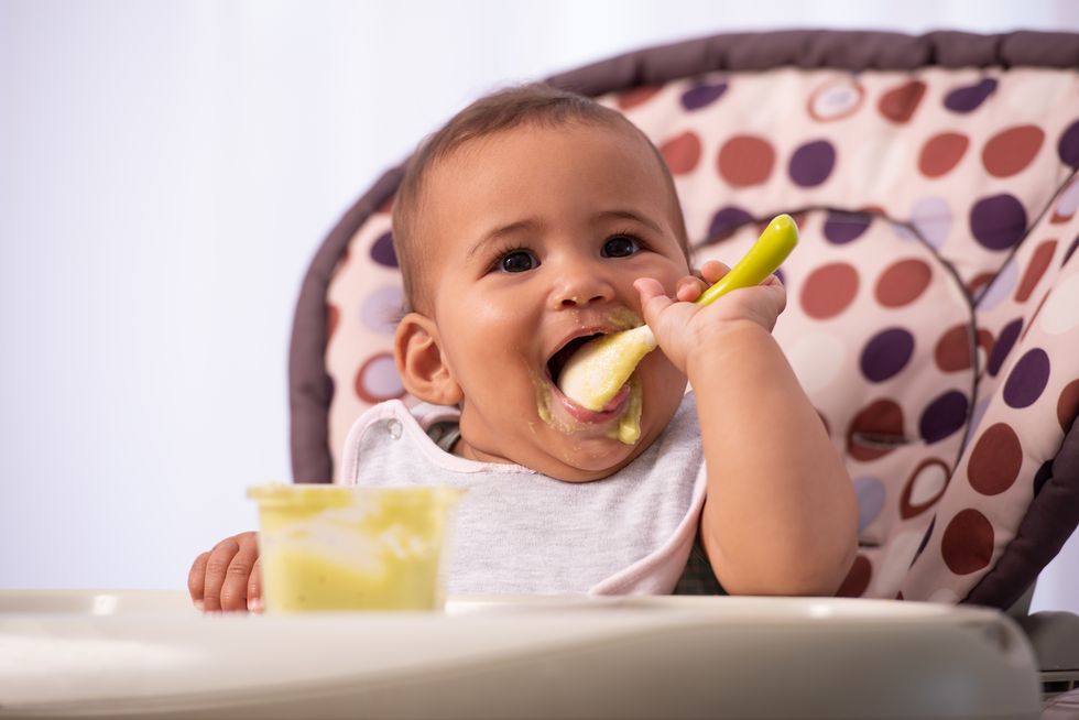 bebé comiendo solo puré de verduras con una cuchara y en una trona