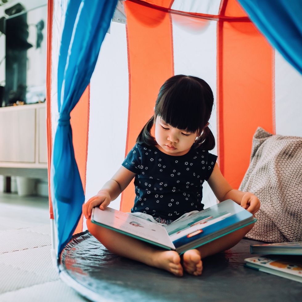 adorable little asian girl enjoying herself and spending leisure time at home she is reading book with her teddy bear in a tent at home