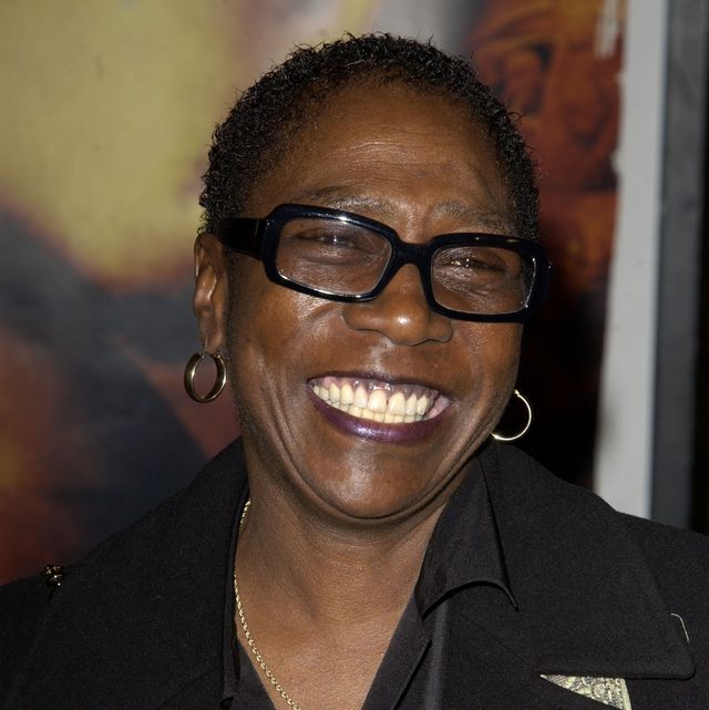 afeni shakure smiles and looks off camera, she is wearing black glasses, hoop earrings and a black outfit