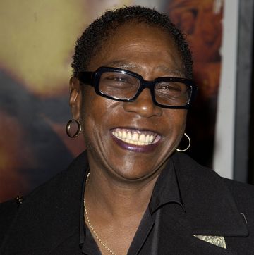 afeni shakure smiles and looks off camera, she is wearing black glasses, hoop earrings and a black outfit