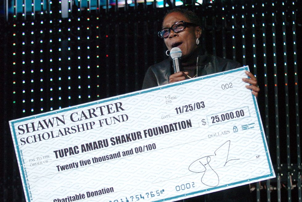 afeni shakur stands on a stand and holds an oversize check in front of her for the tupac amaru shakur foundation, she is also holding a microphone and talking into it, she is wearing black glasses, hoop earrings, and a black outfit
