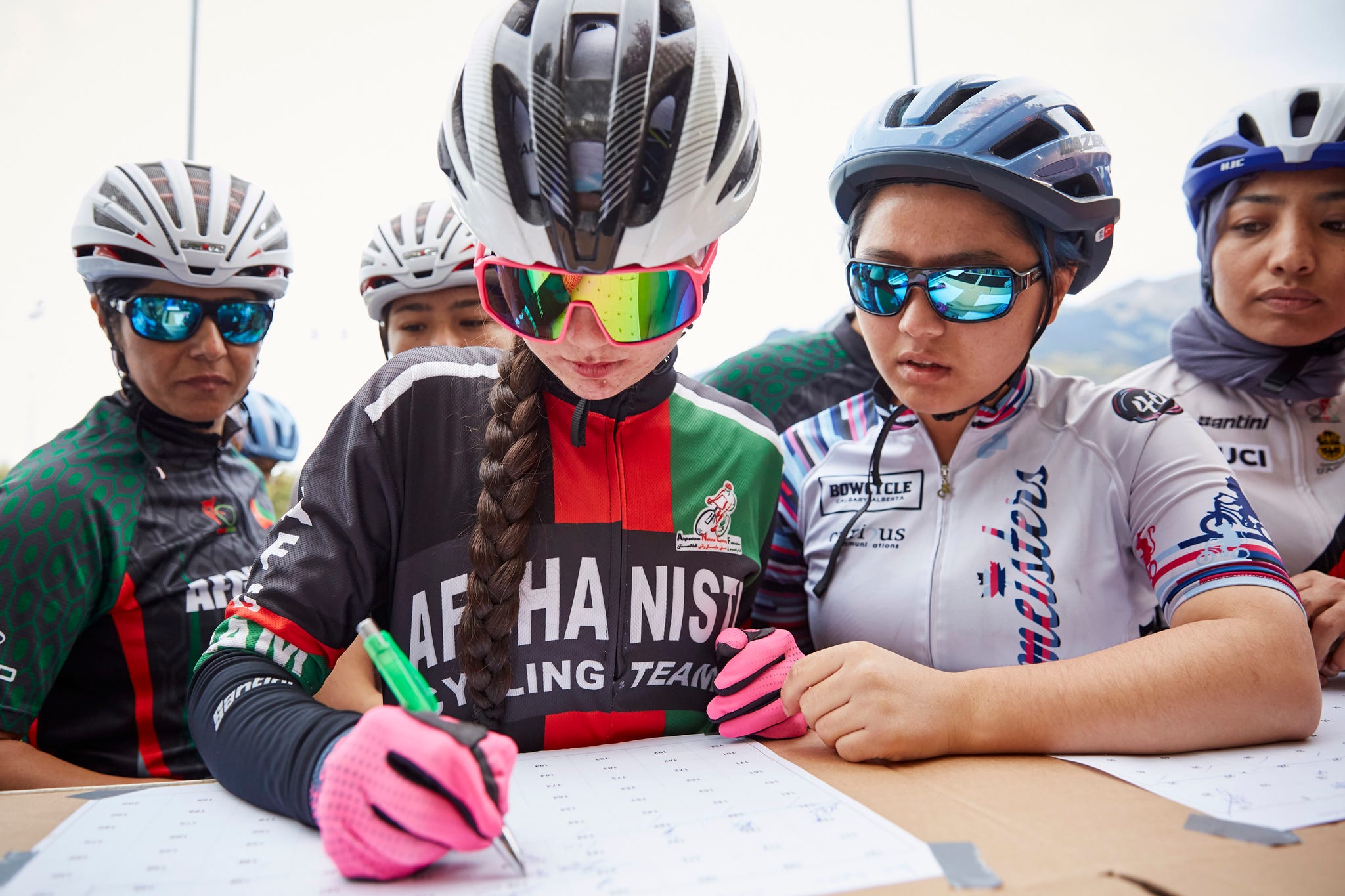 a woman cyclists wearing a helmet and sunglasses signs her name on a sheet surrounded by other cyclists