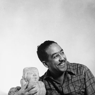 langston hughes smiles and looks right while leaning against a desk and holding a statue sitting on it, he wears a plaid shirt and pants