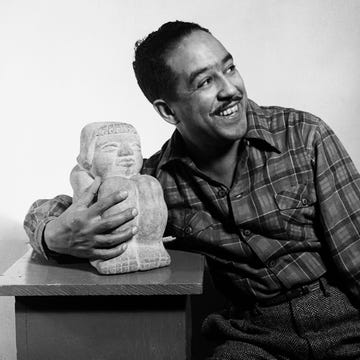 langston hughes smiles and looks right while leaning against a desk and holding a statue sitting on it, he wears a plaid shirt and pants