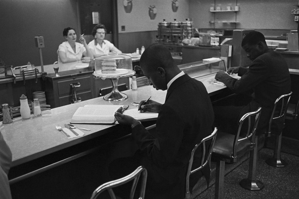 a black and white photo of two young black men sitting at a lunch counter in a restaurant, doing homework while, two white waitresses watch them from the other side of the counter