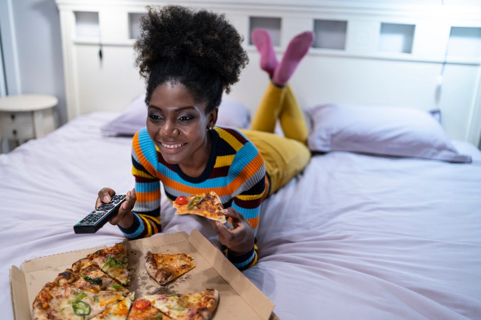 daylight saving sleep - African woman lying down on bed and eating pizza