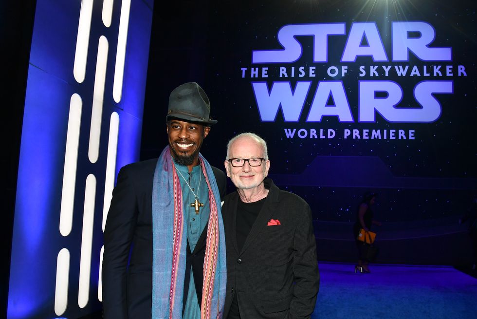 ahmed best, wearing a black suit jacket, black hat, blue shirt, and large blue scarf, stands next to ian mcdiarmid, wearing a black suit jacket and black shirt, in front of a movie screen with the star wars logo