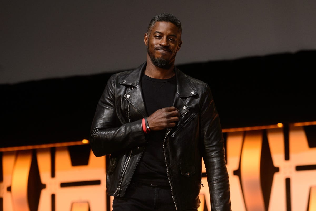 ahmed best wearing a black leather jacket and black shit, holding his hand on his chest and smiling