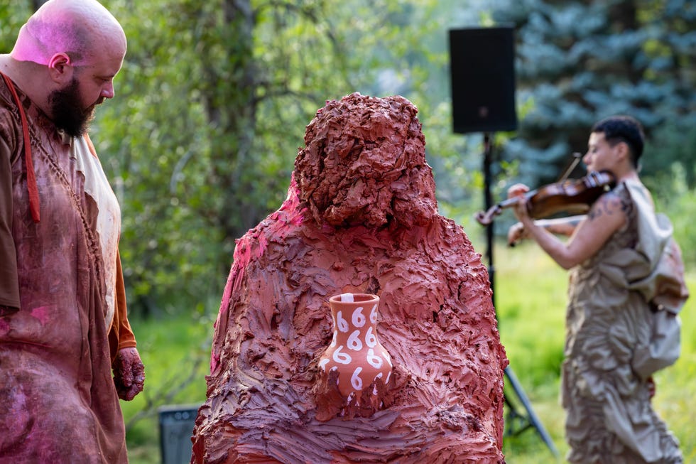 man covered in red clay holding pottery