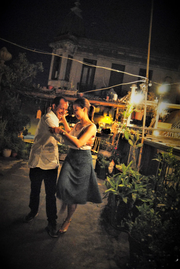 smiling man and woman in a gray skirt tangoing at night in an alley, beside potted plants and a deck
