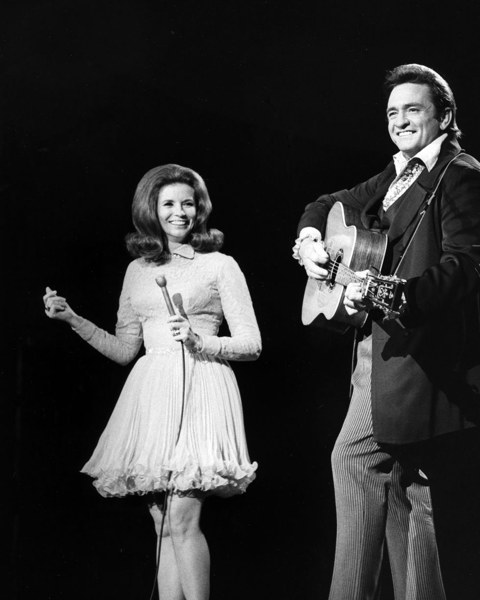 june carter cash and johnny cash stand next to each other and smile, she holds a microphone, he plays a guitar