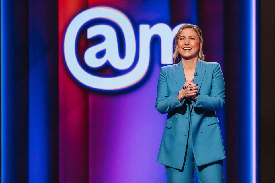 taylor tomlinson smiles while clasping her hands together in front of her chest, she stands in front of a multicolor background with the logo at sign am and wears a bright blue monochrome suit