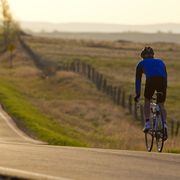 a male cyclist wearing a jacket and shorts, riding along a long undulating road in the early morning light