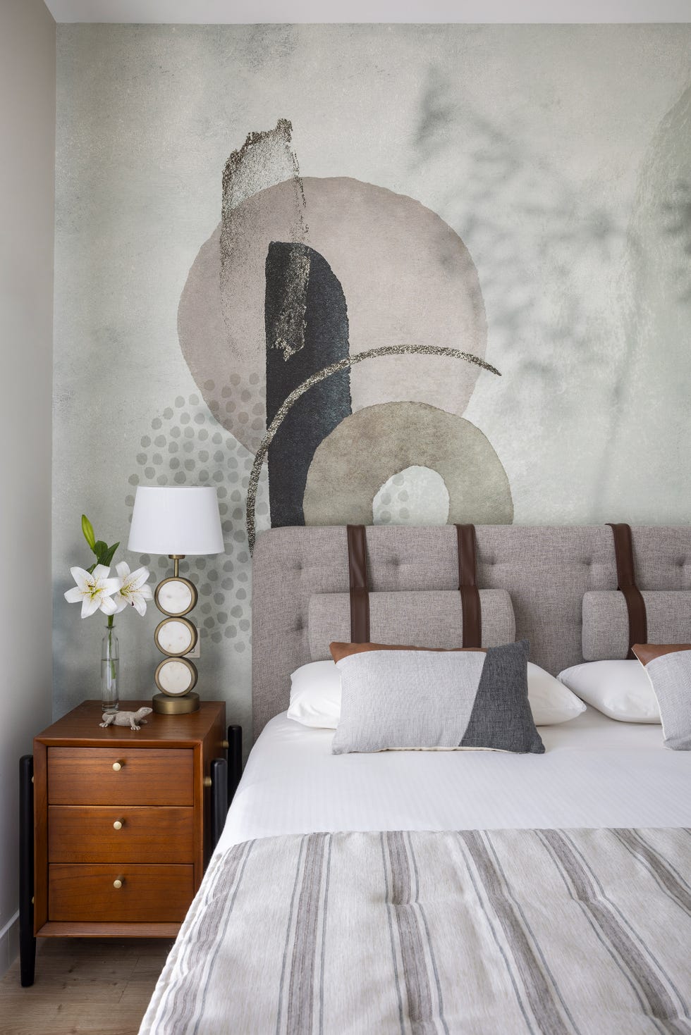 cabecero de cama tapizado en gris con tiras de cuerpo, mesita de madera y papel pintado con motivos abstractos