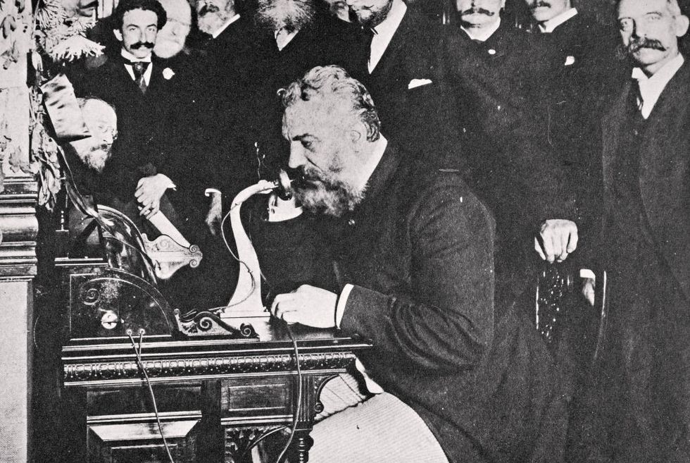 alexander graham bell sits at a desk and holds a phone receiver to his face as he stares forward