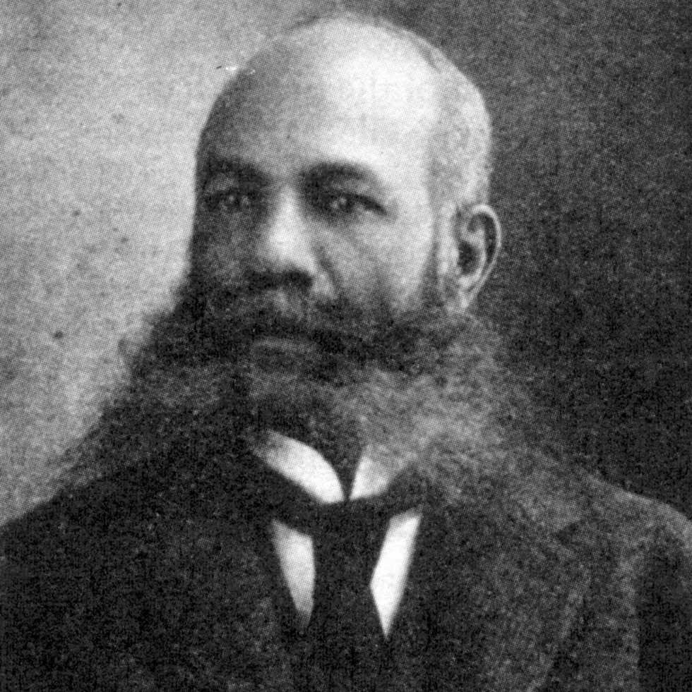 alexander miles looks to the left, he wears a suit jacket, collared shirt and tie with a large full beard