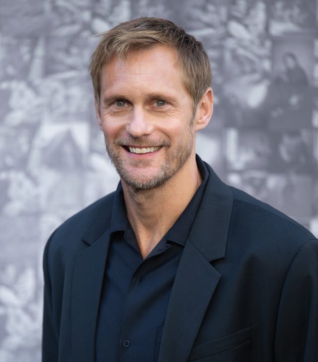 alexander skarsgard smiles at the camera, he wears a dark suit jacket and dark collared shirt