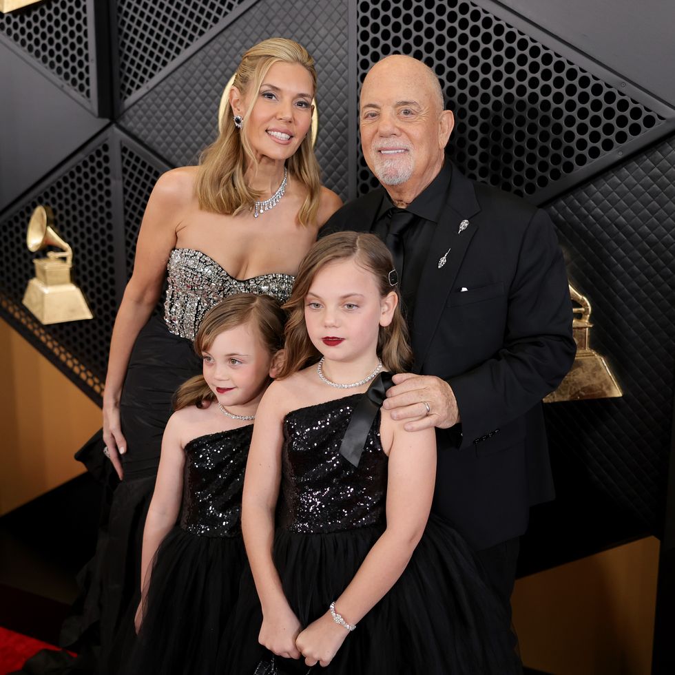 a woman, man and two young girls pose for photos on a red carpet, the woman and girls wear sparkly dresses, the man wears a black suit and tie