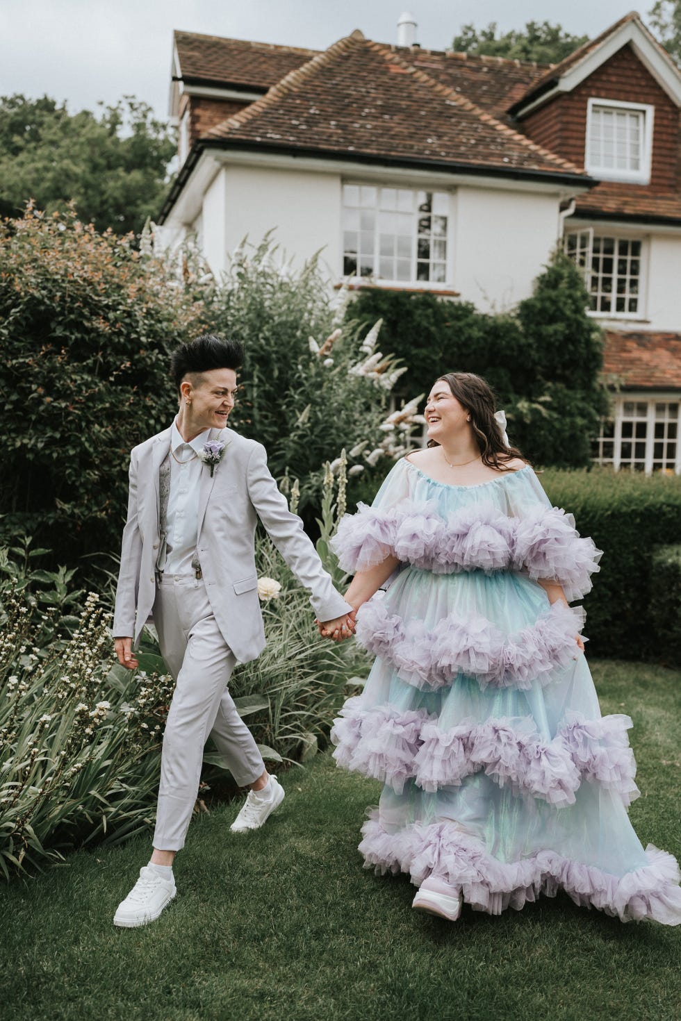 a boy dressed in a light gray suit with a white undershirt and sneakers walks alongside a girl wearing a tiered dress in pastel colors, featuring ruffled layers the background consists of a well kept garden with various plants and a house with a tiled roof visible