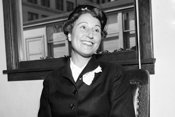 margaret herrick wearing a black dress and hat, sits in a wooden chair and smiles while sitting in front of a window